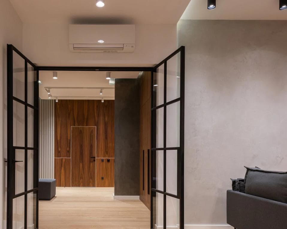 Modern interior of a hallway with wooden door and black metal framed glass doors, featuring recessed lighting and a sleek air conditioning unit