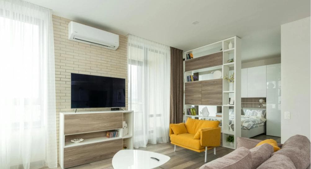 Bright living space with a yellow sofa, white curtains, and a partition shelf unit, complemented by a wall-mounted air conditioner